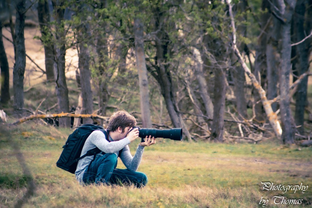 Thomas photographing with a telephoto lens in a field at the edge of a forest